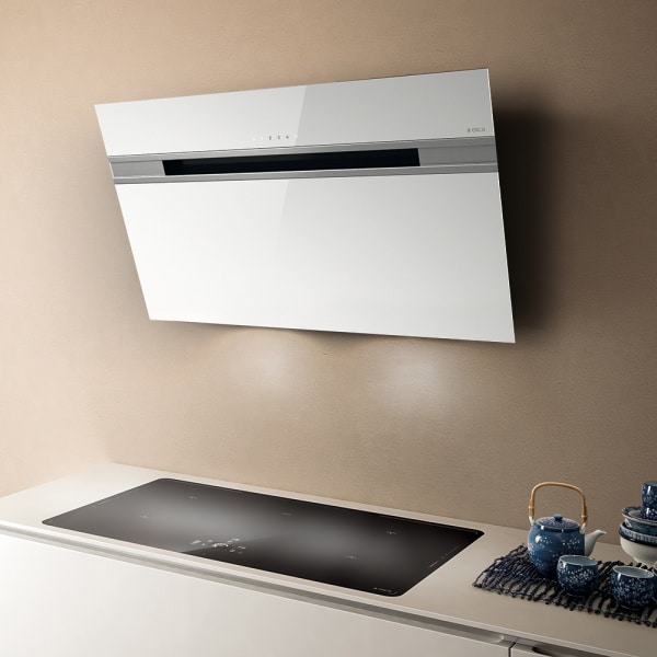 Na obrázku je kuchynský digestor zobrazený z boku, namontovaný na stene nad varnou doskou. Digestor má moderný dizajn s hladkým povrchom a je bielej farby s lesklým povrchom. Na prednej strane sú viditeľné dotykové ovládacie prvky a jemné nápisy. Pod digestorom je čierna varná doska, ktorá je zabudovaná do bielej kuchynskej linky. Na linke sú umiestnené modré keramické čajové súpravy. Prostredie je interiérové, pravdepodobne kuchyňa, a osvetlenie je denné alebo neutrálne. Zdôraznený je elegantný a čistý dizajn kuchynského vybavenia.