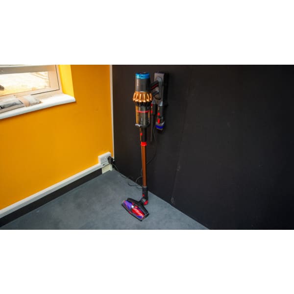 Na obrázku je bezdrôtový tyčový vysávač zobrazený z boku, umiestnený na nabíjacej stanici pripevnenej na stene. Vysávač má kovový rám v bronzovej farbe s lesklým povrchom. Zberná nádoba je priehľadná, čo umožňuje vidieť jej obsah. Hlava vysávača je vybavená rotačnou kefou s farebnými prvkami, najmä červenými a modrými. Na stene je viditeľná nabíjacia stanica, ku ktorej je vysávač pripojený káblom. Prostredie je interiérové, s oranžovou stenou a oknom, cez ktoré preniká denné svetlo. Na vysávači sú viditeľné nápisy, ktoré označujú značku alebo model.